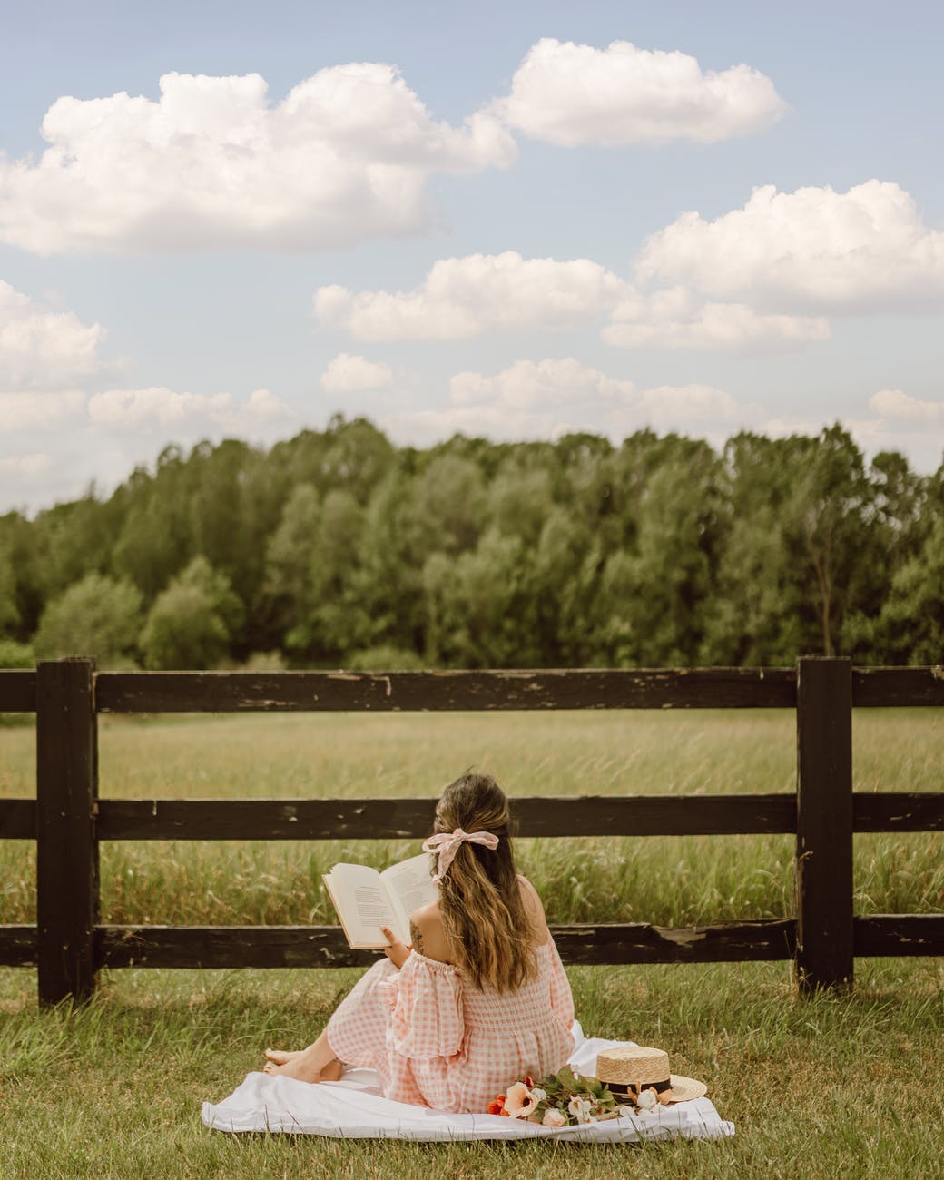 back view of a woman reading a book while sitting on a picnic blanket near the wooden fence for how reading can improve your mental health post.
