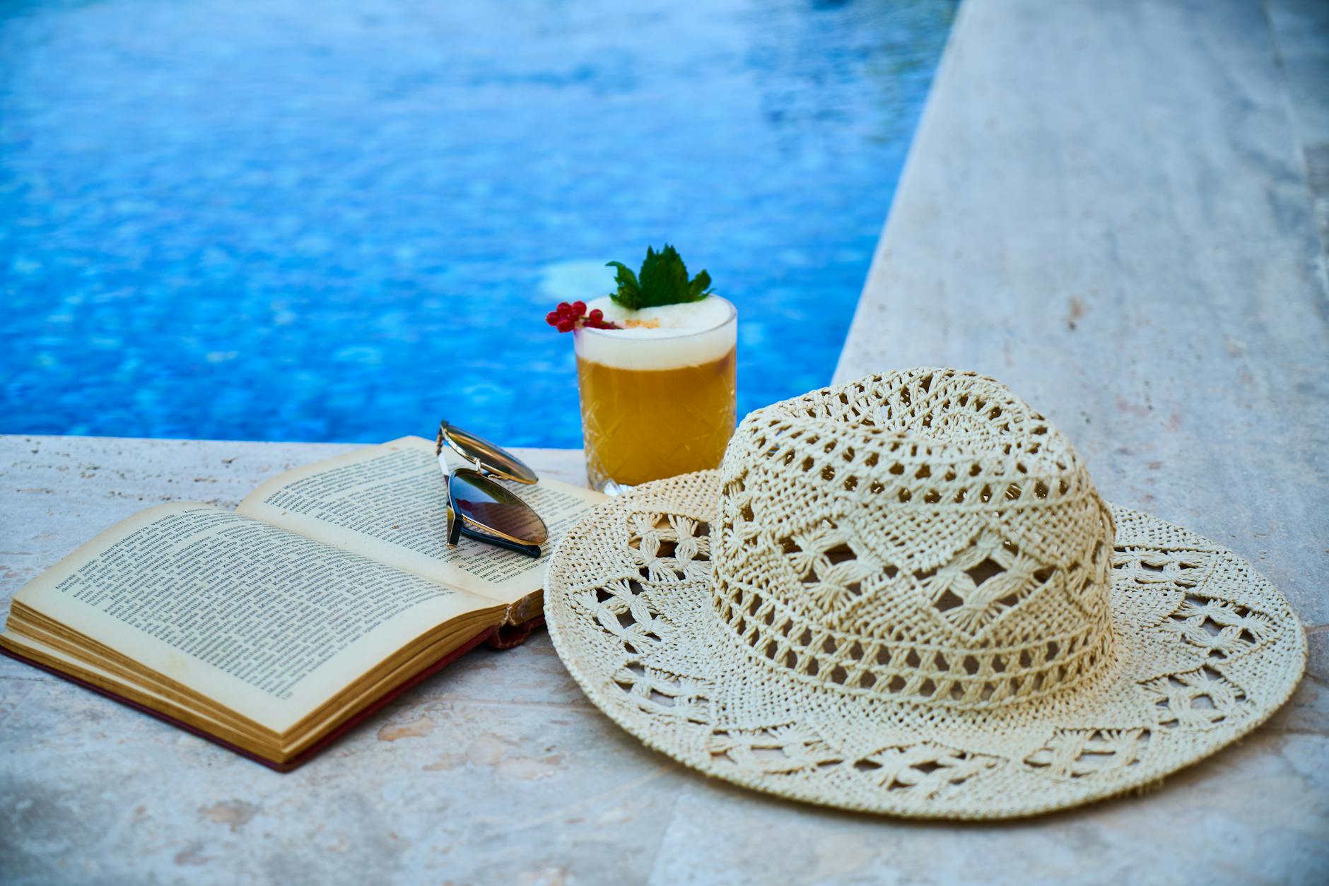 Hat, cocktail, sunglasses and book poolside for travel thriller books to add to your packing list post. 