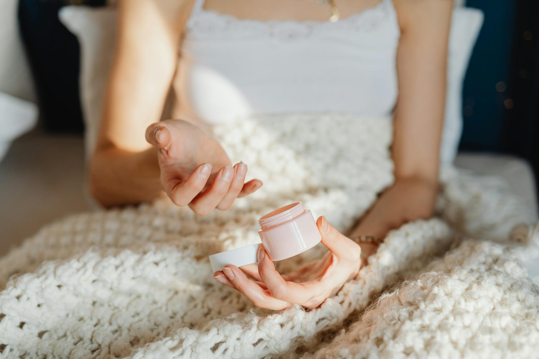 Person under a blanket getting face cream out of a pot for Winter Skincare Tips to Combat Dryness and Cold post.